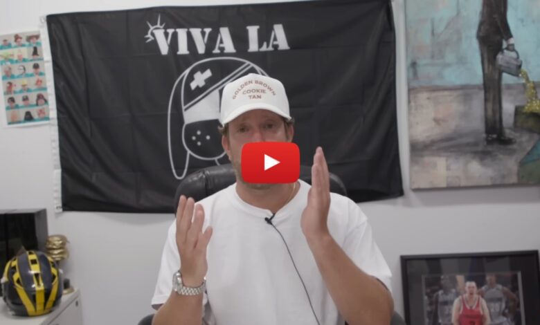 Screenshot from Dave Portnoy's YouTube video showing him wearing a white 'Golden Brown Cookie Tan' cap and white shirt speaking on camera in front of a 'Viva La' flag, with sports memorabilia and art in the background.