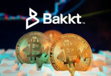 Three Bitcoin on computer keyboard, with white Bakkt logo on dark blue stock chart background.