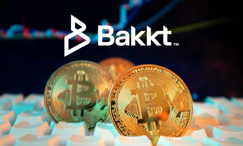 Three Bitcoin on computer keyboard, with white Bakkt logo on dark blue stock chart background.