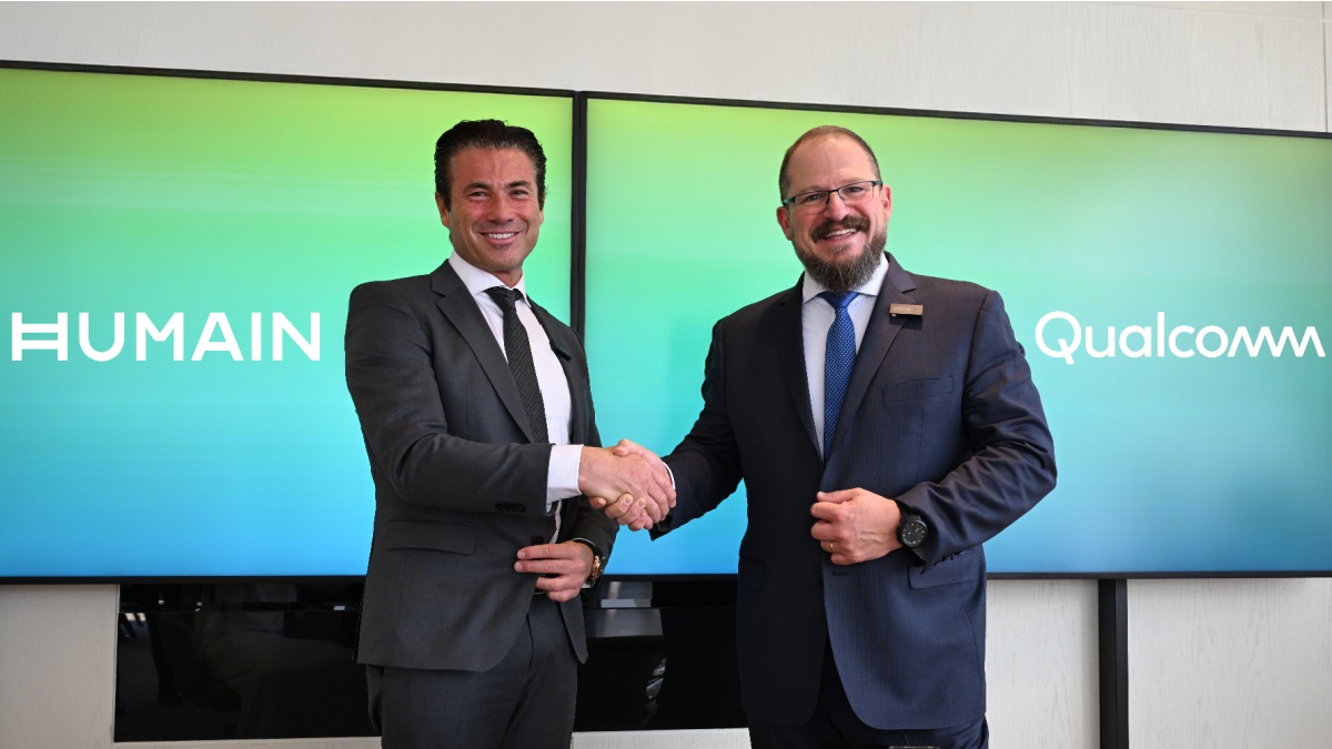 Qualcomm and HUMAIN top executives shake hands in front of a green screen with company logos.