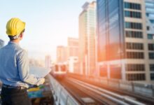 Construction worker wearing a yellow hard hat and holding blueprints, standing on an elevated platform overlooking a train track and modern city skyline with high-rise buildings during sunset.