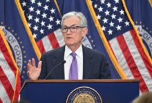 Federal Reserve Chair Jerome Powell in a suit and purple tie speaks at a podium with the seal of the Board of Governors in front. He is gesturing with one hand and standing in front of several American flags and blue banners with gold trim.