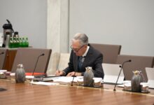 Federal Reserve Chair Jerome Powell sits alone at a conference table, writing on documents. Several microphones, pitchers, and tablets are arranged on the table. In the background, a counter holds bottled water, coffee supplies, and a security camera is mounted on the wall.