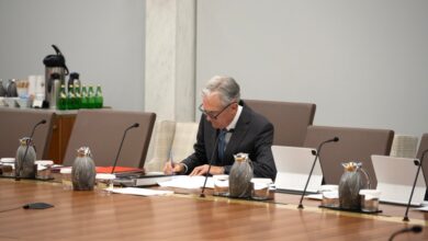 Federal Reserve Chair Jerome Powell sits alone at a conference table, writing on documents. Several microphones, pitchers, and tablets are arranged on the table. In the background, a counter holds bottled water, coffee supplies, and a security camera is mounted on the wall.