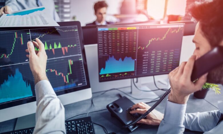 Stock trader on the phone pointing at charts on monitors, with financial graphs and data visible in a busy office.