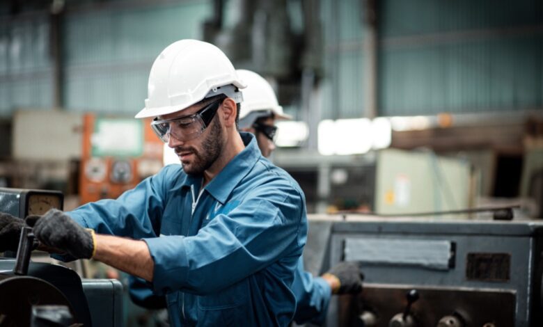 Factory worker in a hard hat and safety glasses operating industrial machinery in a workshop.