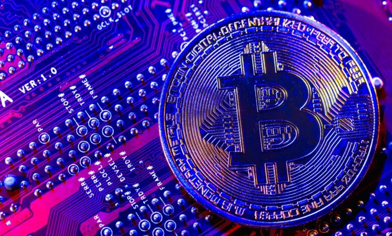 Close-up of a metallic bitcoin coin resting on a colorful computer circuit board with glowing blue and purple tones.