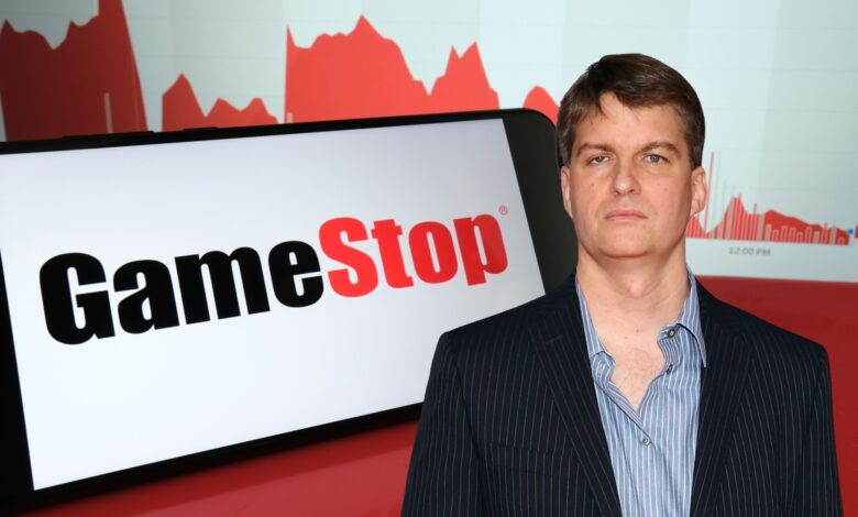 Michael Burry in a pinstripe suit standing in front of a large GameStop logo and red stock chart graphics.
