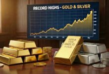 Stacks of gold and silver bars with coins on a wooden table in front of a chart showing record highs in gold and silver prices.