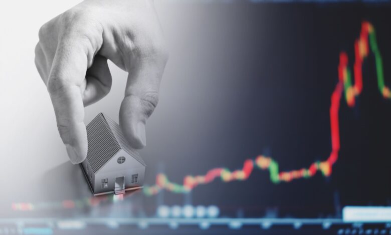 A hand places a small model house in front of a blurry background showing a red and green candlestick chart, symbolizing real estate investment or housing market trends.
