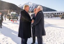President Donald Trump shaking hands with a man in a snowy mountain setting in Davos, Switzerland, surrounded by officials and helicopters.