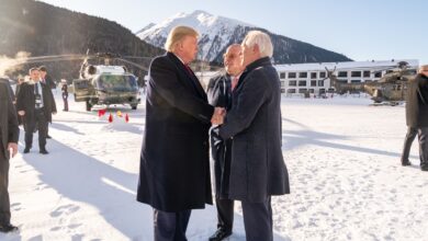 President Donald Trump shaking hands with a man in a snowy mountain setting in Davos, Switzerland, surrounded by officials and helicopters.