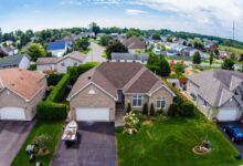Aerial view of a suburban neighborhood with rows of single-family homes, green lawns, and a playground in the background under a partly cloudy sky.