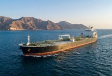 Aerial wide shot of a large black oil tanker cargo ship cruising on calm blue sea near rugged rocky coastal mountains at golden hour, with clear sky, ship deck piping and superstructure visible, maritime transportation, marine logistics, petroleum shipping, ocean freight vessel.