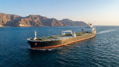 Aerial wide shot of a large black oil tanker cargo ship cruising on calm blue sea near rugged rocky coastal mountains at golden hour, with clear sky, ship deck piping and superstructure visible, maritime transportation, marine logistics, petroleum shipping, ocean freight vessel.