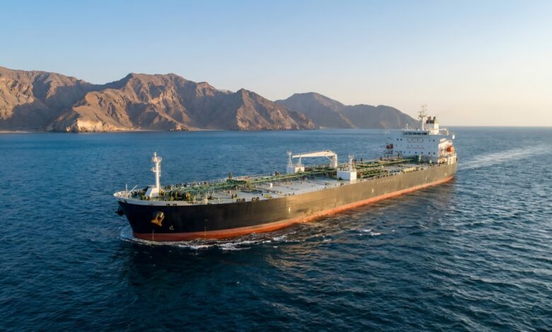 Aerial wide shot of a large black oil tanker cargo ship cruising on calm blue sea near rugged rocky coastal mountains at golden hour, with clear sky, ship deck piping and superstructure visible, maritime transportation, marine logistics, petroleum shipping, ocean freight vessel.